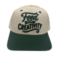 A two-tone baseball cap with a beige crown and a dark green flat brim, featuring the phrase 'Feed YOUR CREATIVITY' embroidered on the front in dark green.
